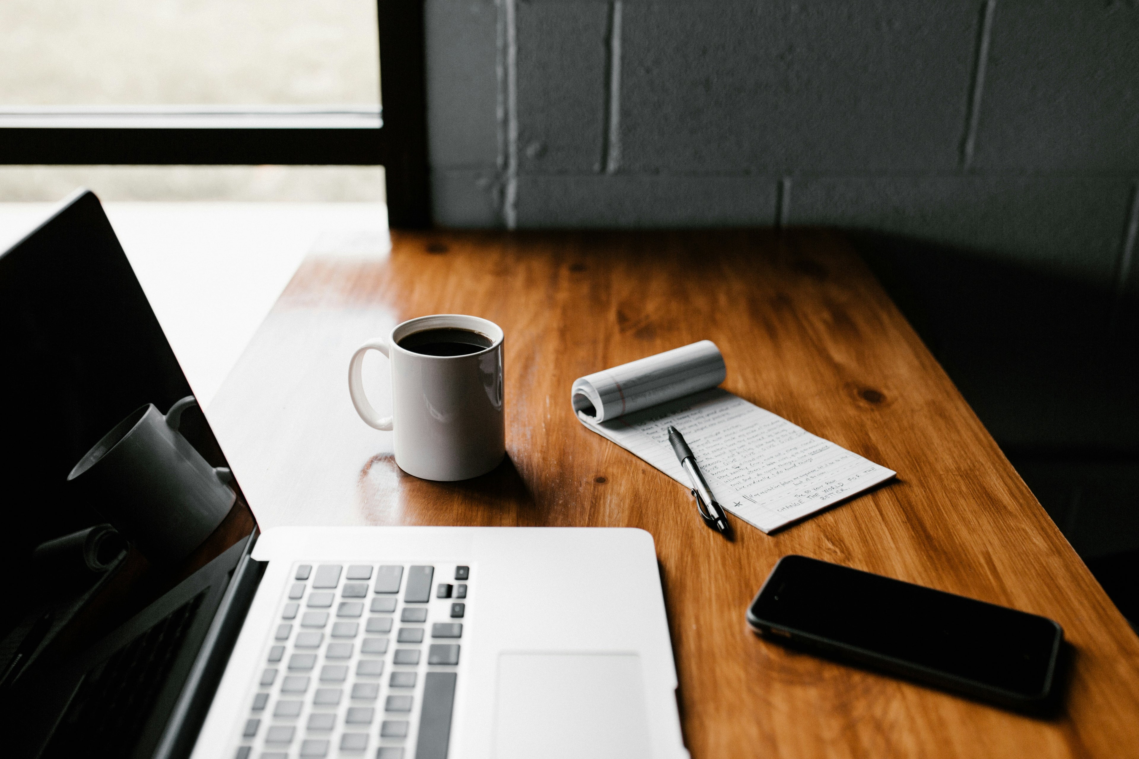 Laptop, coffee mug, notebook, pen, and smartphone on a wooden table.